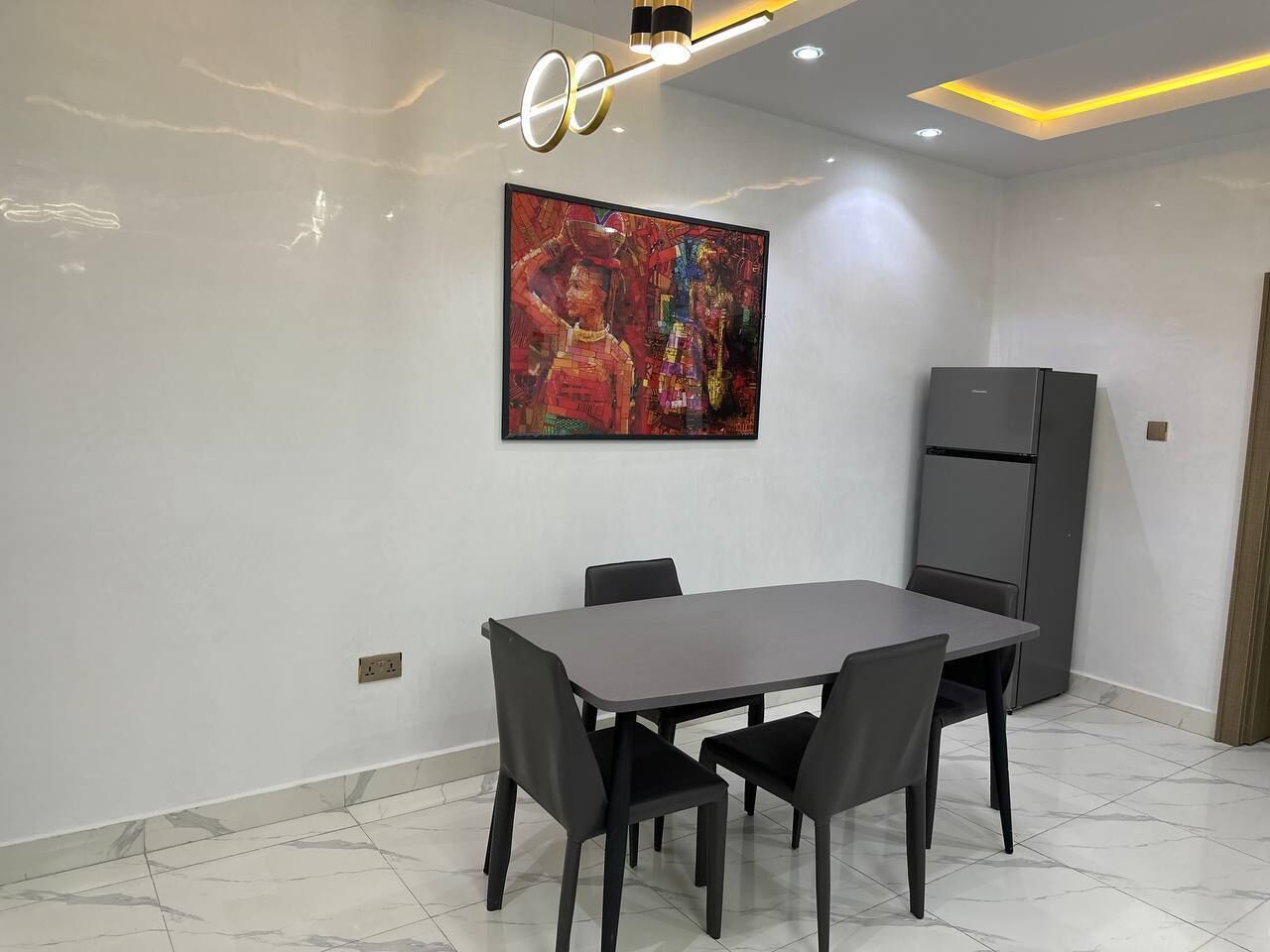 A modern dining area with a rectangular table and four chairs, a large abstract painting on the wall, a refrigerator, and a recessed ceiling light.