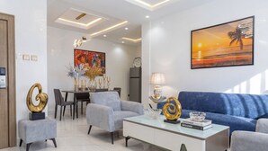 A modern living room with a blue sofa, grey armchairs, a glass coffee table, and a dining area with a chandelier. - Prestine Apartments (Abuja)