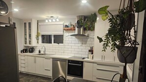 A modern kitchen with white cabinets, a stainless steel refrigerator, a dishwasher, and a sink. There are hanging plants and a window with blinds.