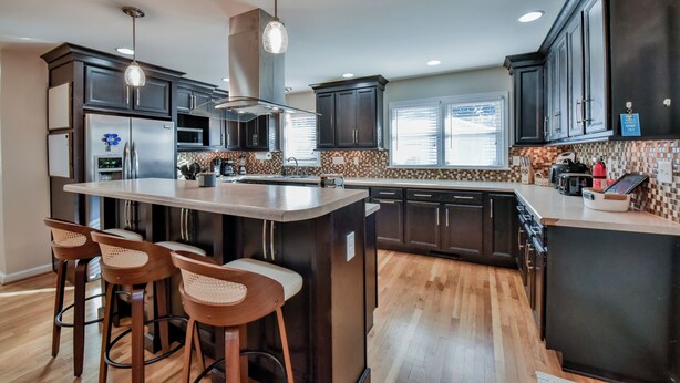 A modern kitchen with dark cabinetry, a central island with bar stools, stainless steel appliances, and a patterned backsplash. - TuscaLodge: Close to Stadium, Big on Comfort! (Tuscaloosa)