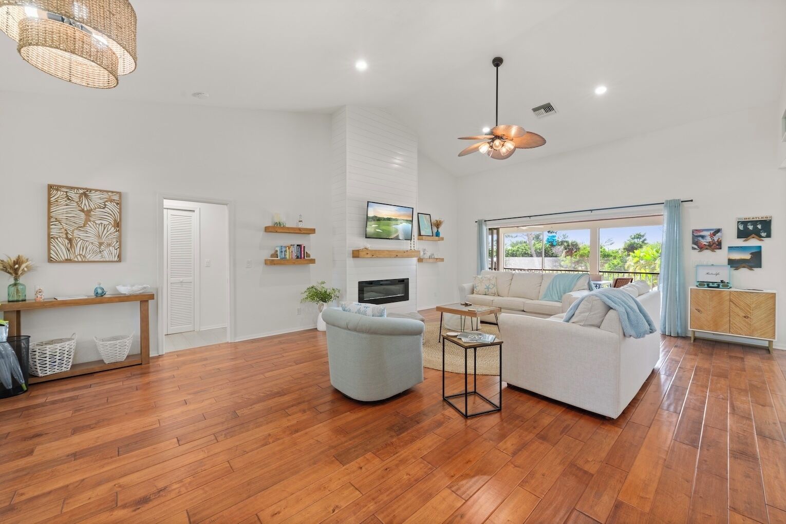 A spacious living area with a large sofa, a coffee table, and a television. There are wooden floors, a ceiling fan, and a window with a view of greenery.