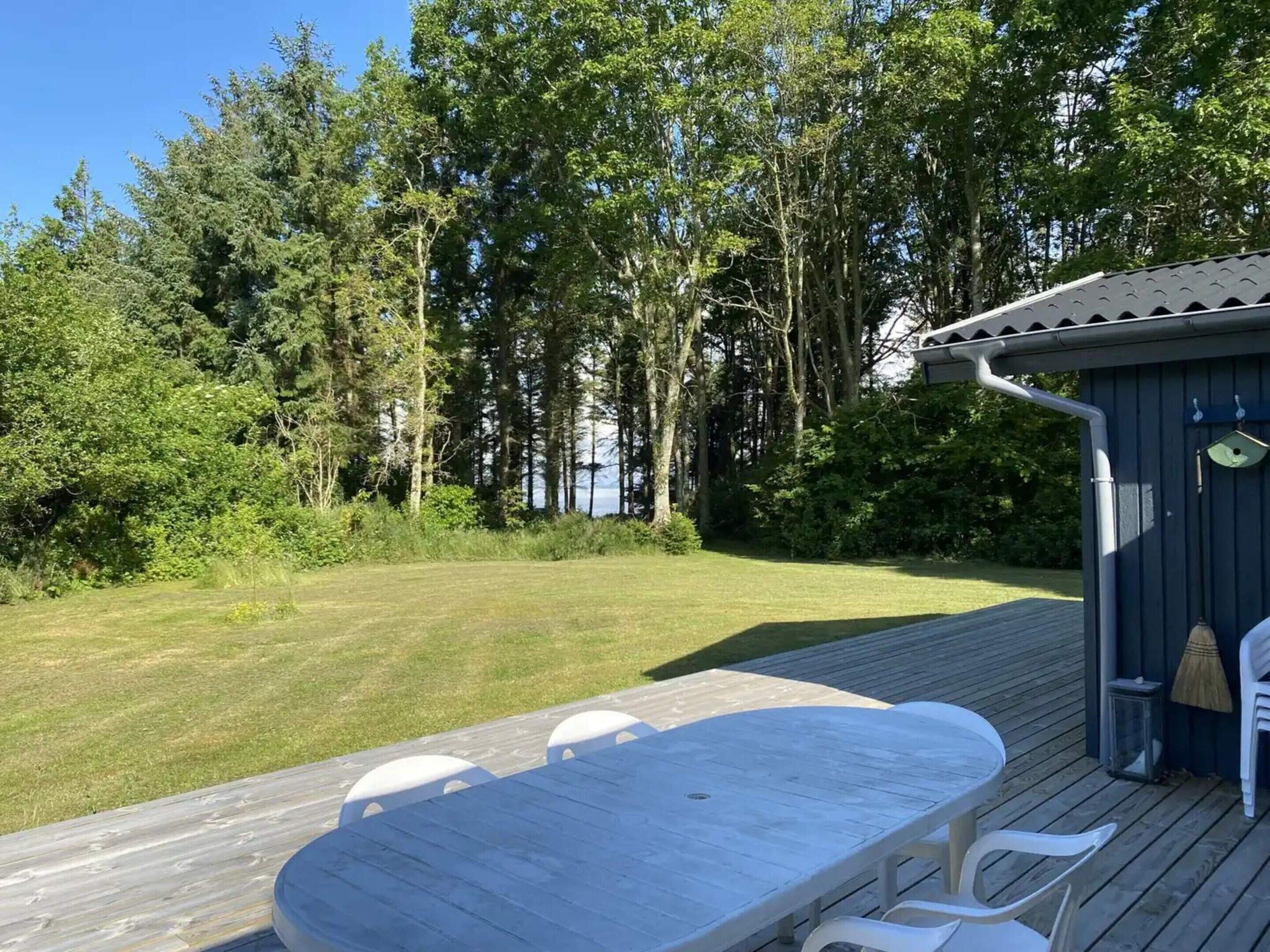 A wooden deck with a table and chairs, surrounded by a grassy area and dense trees.