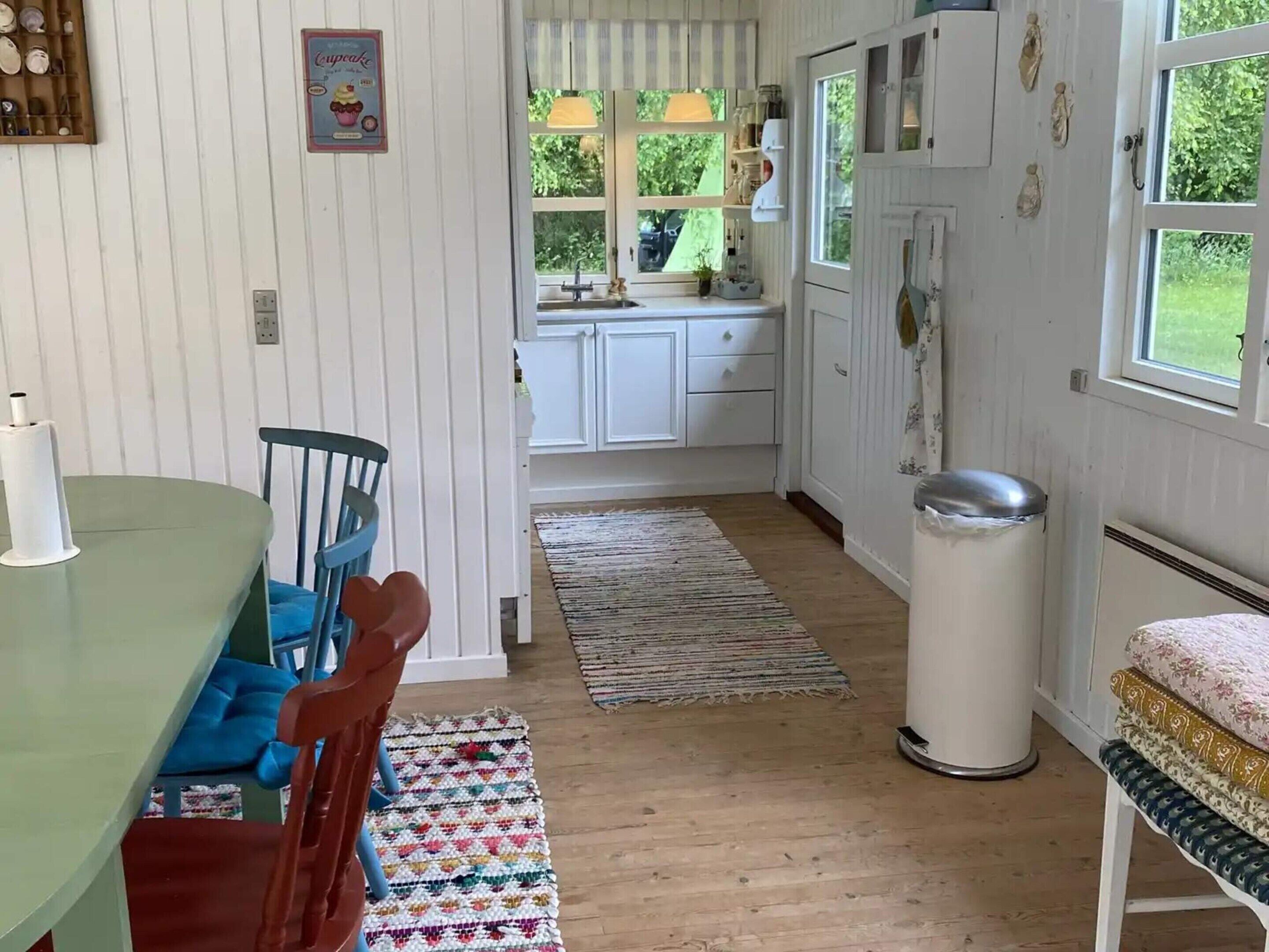 Una cocina acogedora con armarios blancos, una mesa de comedor con sillas, un cubo de basura y una ventana con vista a zonas verdes.