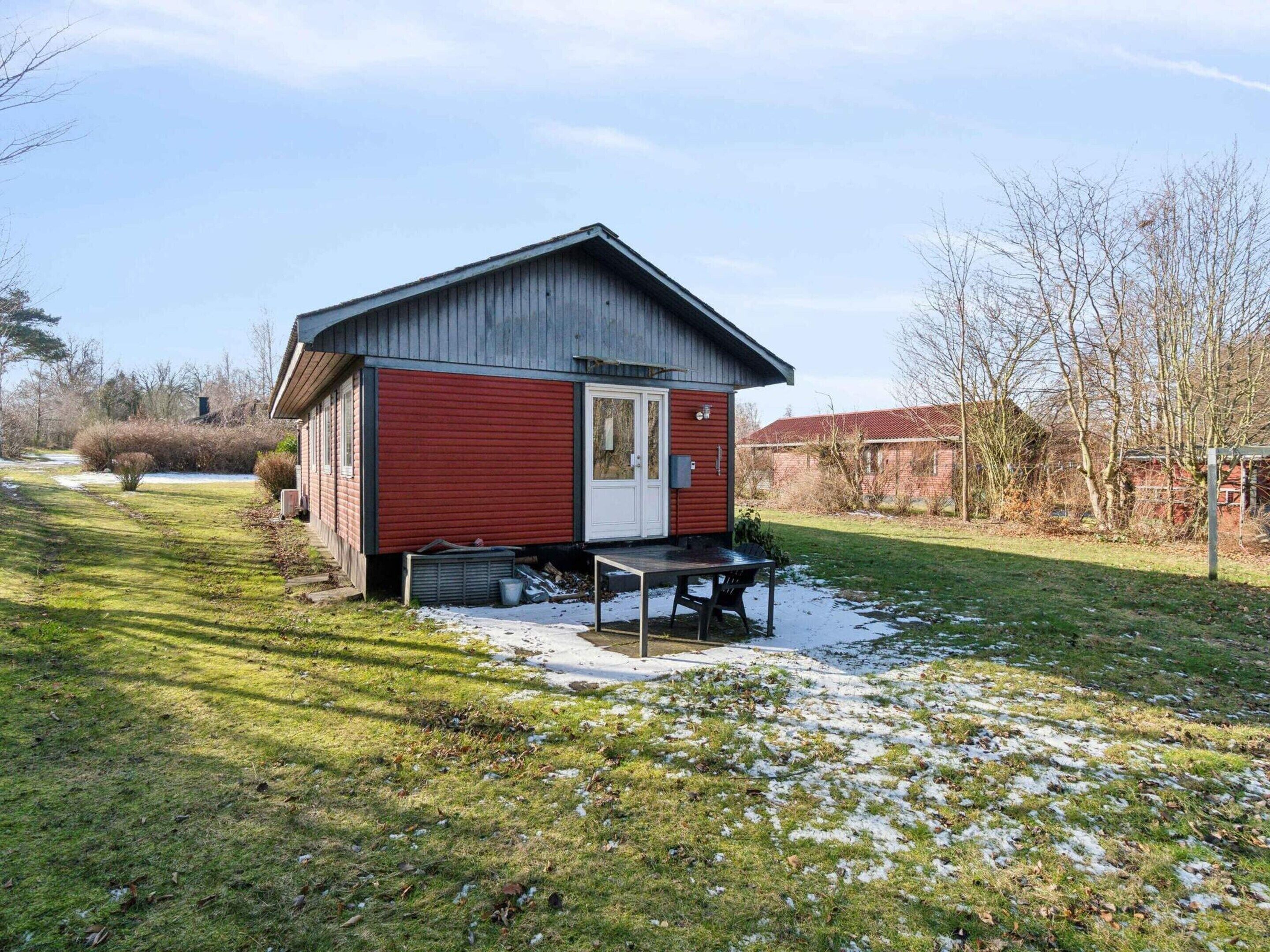 A small, single-story building with a red exterior and a white door, situated in a grassy area with some snow patches.