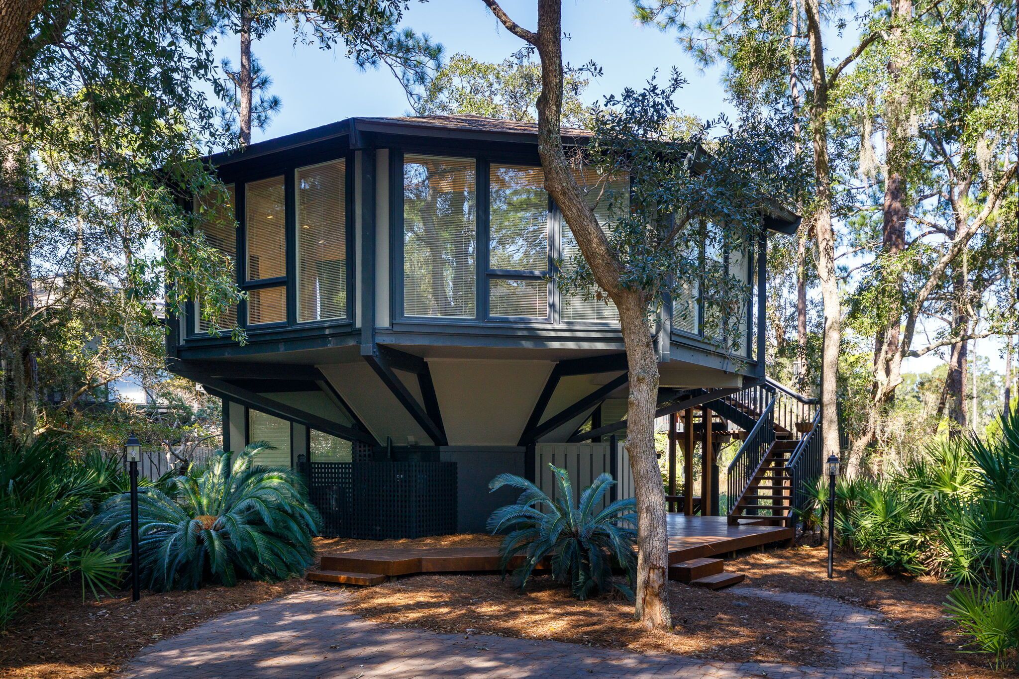 Une maison moderne à plusieurs niveaux, dotée de grandes fenêtres et entourée d’une végétation luxuriante et d’arbres.