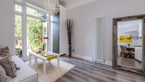 A spacious living room with a white sofa, a small coffee table, and a large mirror. There is a window with a view of greenery outside. - Elegant Ealing Apartment Retreat (Cape Town)
