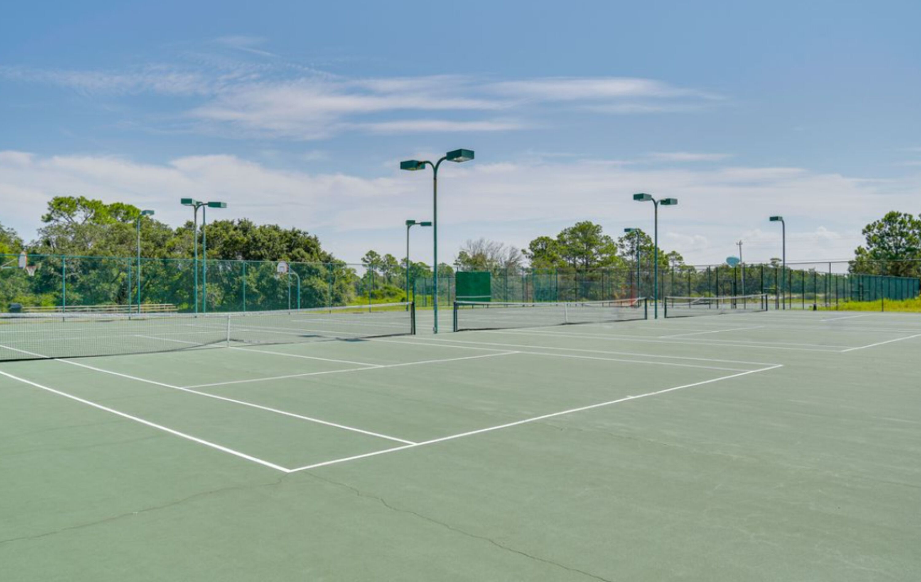 An outdoor tennis court with green flooring, surrounded by a fence, and street lamps.