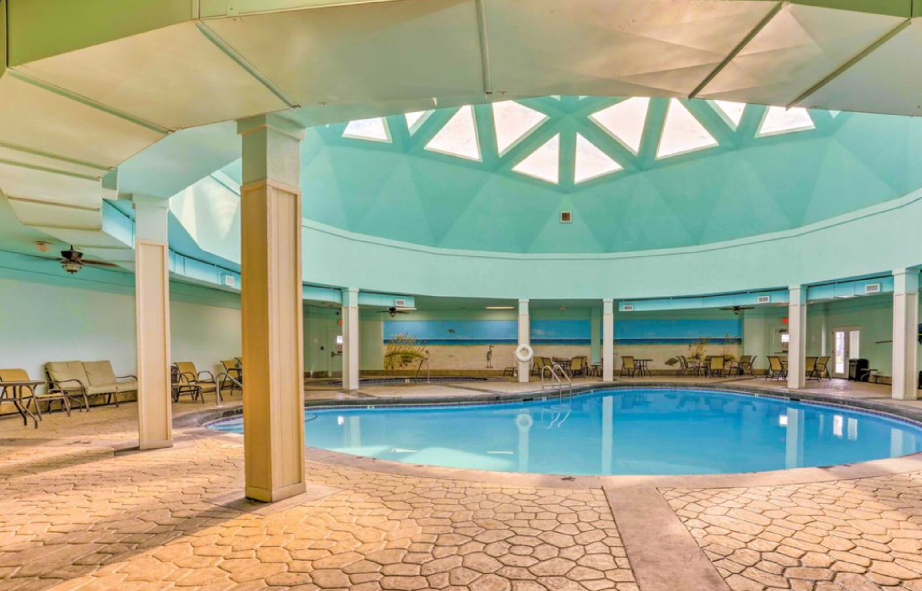 An indoor swimming pool with a tiled floor, lounge chairs, and a high ceiling with skylights.