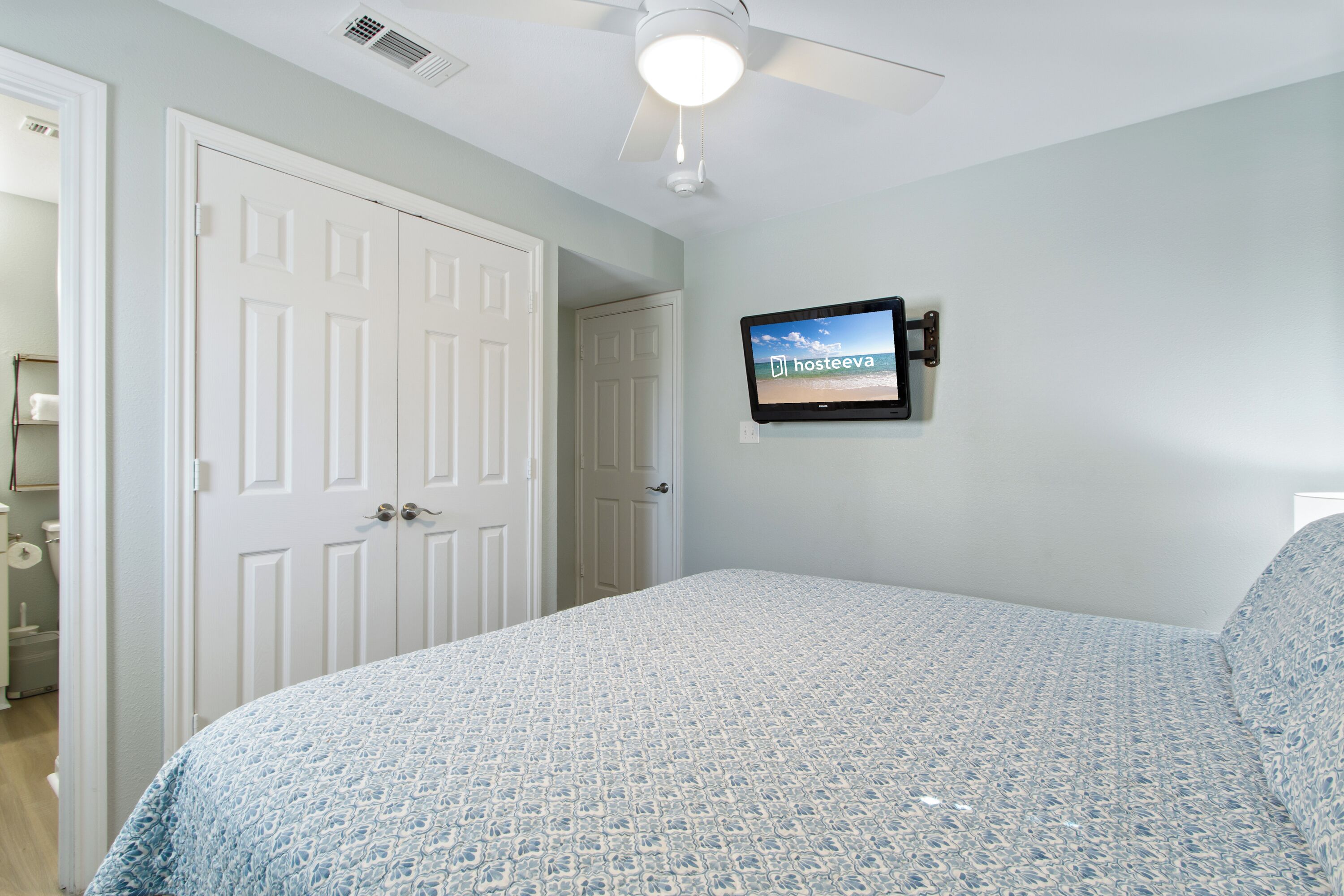 A bedroom with a bed, a ceiling fan, a TV mounted on the wall, and a door leading to another room.