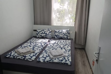A single bed with a patterned bedspread and pillows, a window with curtains, and a wooden floor.