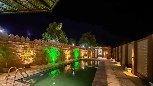 Un espace piscine aménagé pour la nuit, avec un mur en pierre, des plantes en pot et un petit pont enjambant la piscine.