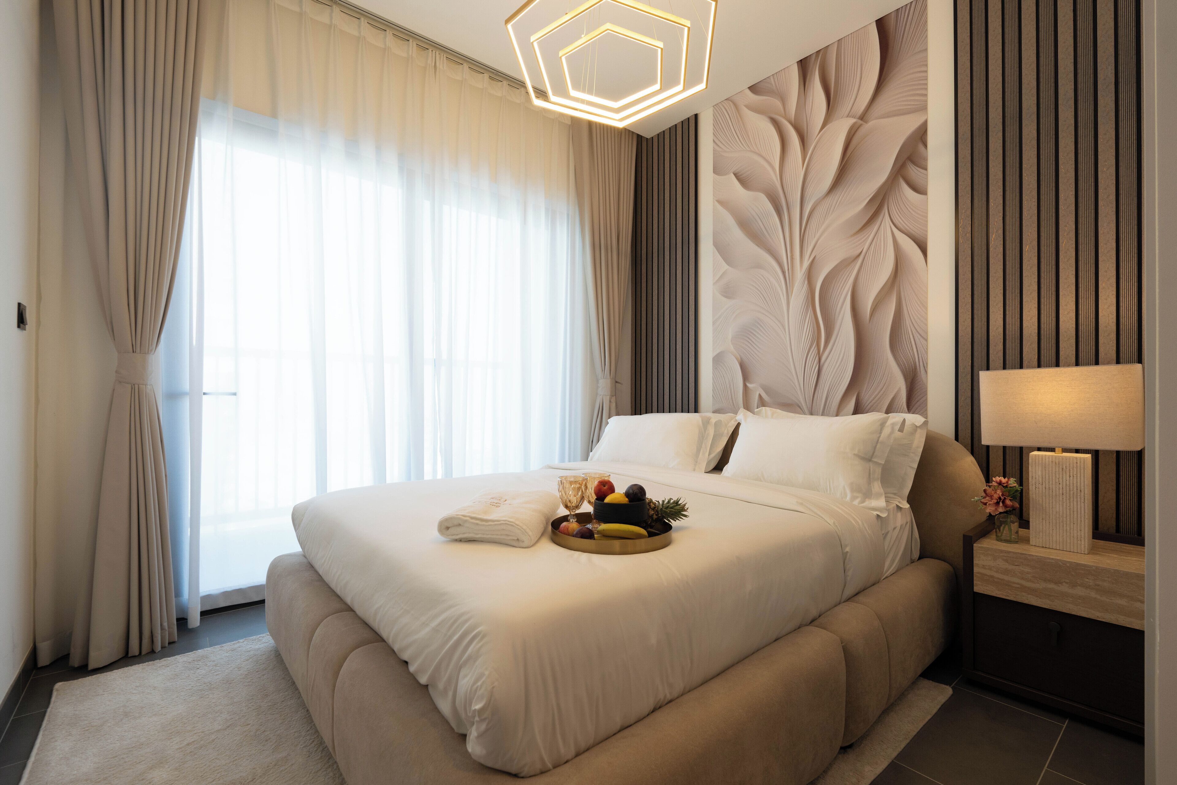 Una habitación de hotel moderna con una cama grande, una mesita de noche con lámpara y una cesta de frutas sobre la cama.