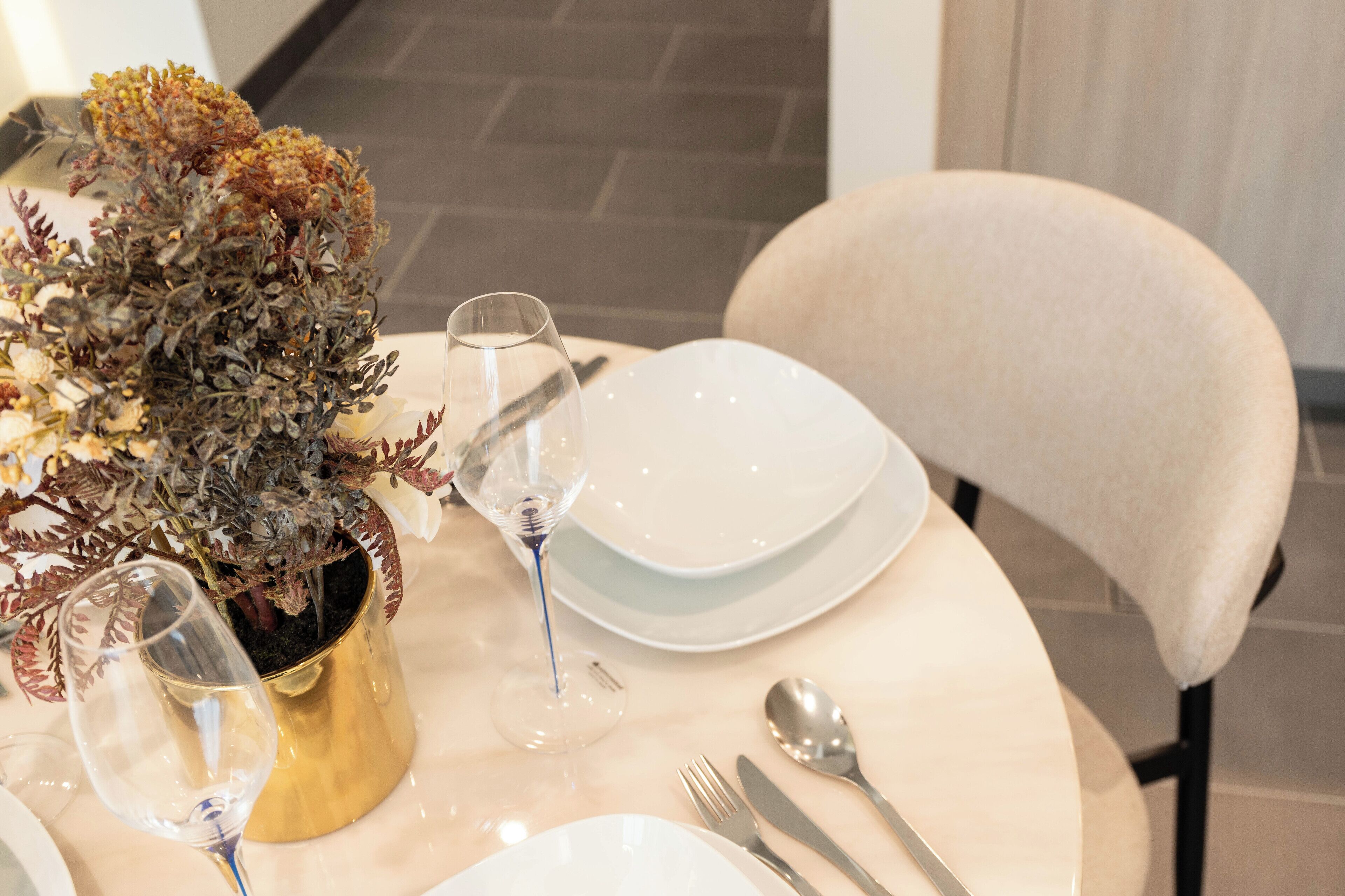 A dining table set with white plates, silverware, and a gold vase with dried flowers.