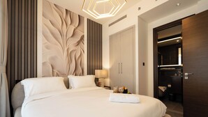 Quarto de hotel moderno com uma cama grande, um painel decorativo na parede, um guarda-roupa de madeira e um banheiro visível por uma porta aberta.