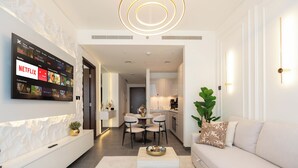 Uma sala de estar moderna com uma TV de tela plana exibindo Netflix, uma área de jantar com mesa e cadeiras redondas, e uma cozinha ao fundo.