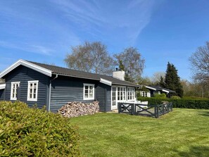 A two-story house with a dark exterior, white trim, and a chimney. There is a stack of firewood in front of the house, and a wooden deck with a railing. - 5 Star Holiday Home in Kirke Hyllinge (Kirke Hyllinge)