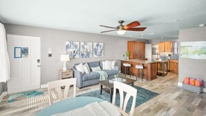 A modern living room with a sofa, coffee table, and chairs, a kitchen area with wooden cabinets, and a ceiling fan. - Private Beach Access Sauna Outdoor Fireplace (St. Augustine)