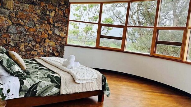 A bedroom with a stone wall, a bed with a tropical leaf-patterned bedspread, and large windows offering a view of trees. - Wandin Farm Views - architecturally designed home (Wandin North)