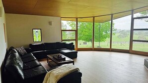 A spacious living room with a large L-shaped black leather sofa, a wooden coffee table, and a view of a green landscape through large windows. - Wandin Farm Views - architecturally designed home (Wandin North)
