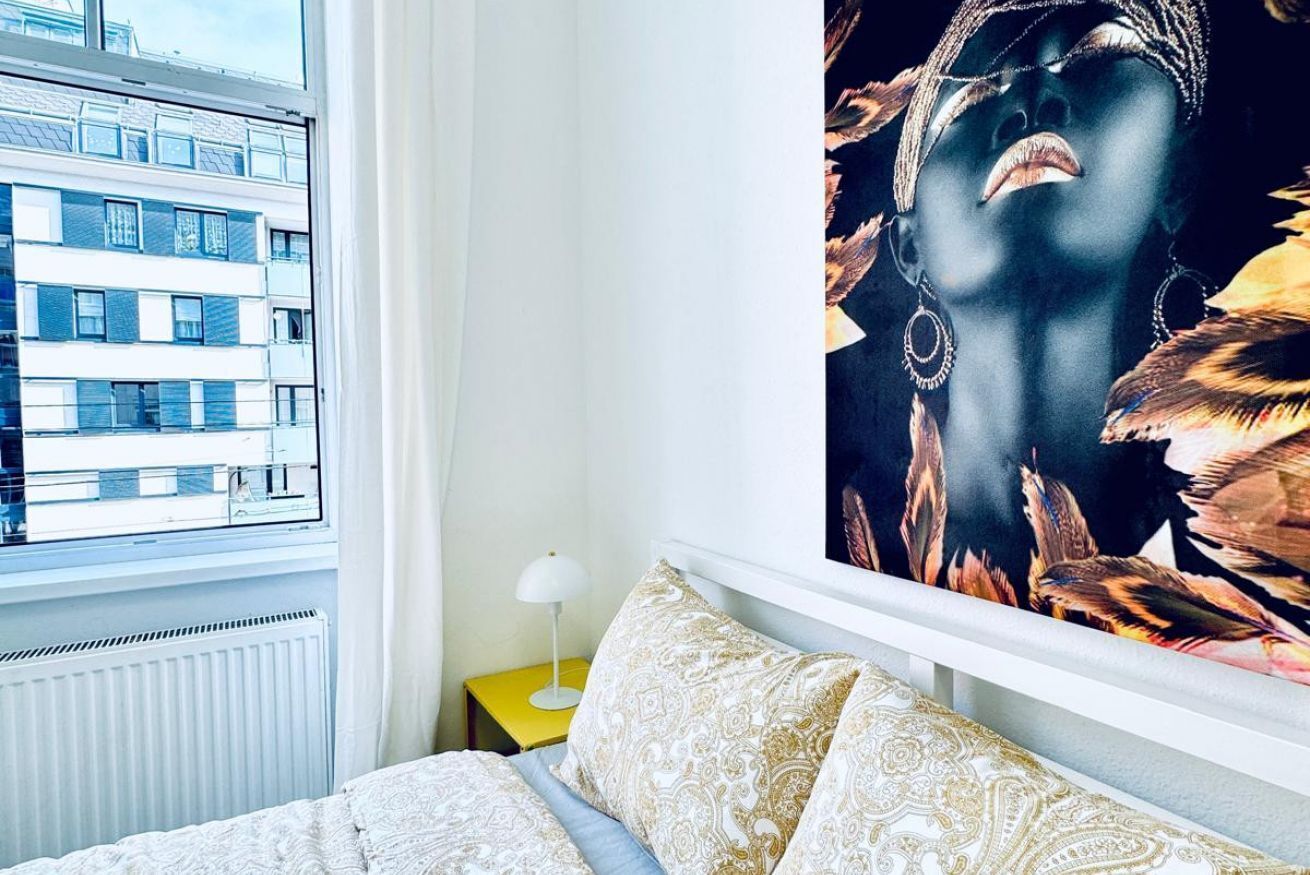 Une chambre à coucher avec un lit, une fenêtre donnant sur des immeubles, une table de chevet jaune avec une lampe, et un grand tableau accroché au mur.