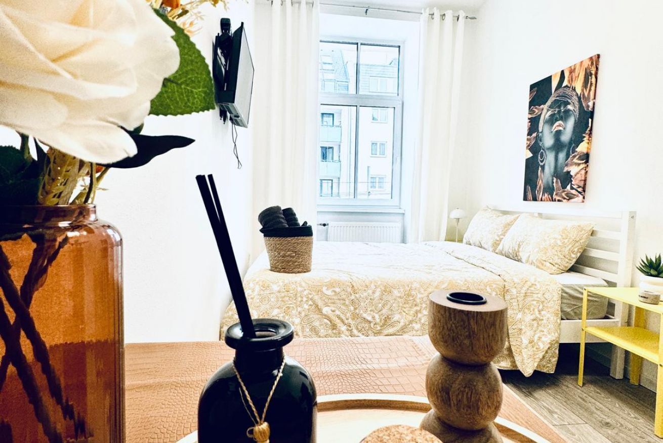 A bedroom with a bed, a window with a view, a vase with flowers, and a painting on the wall.