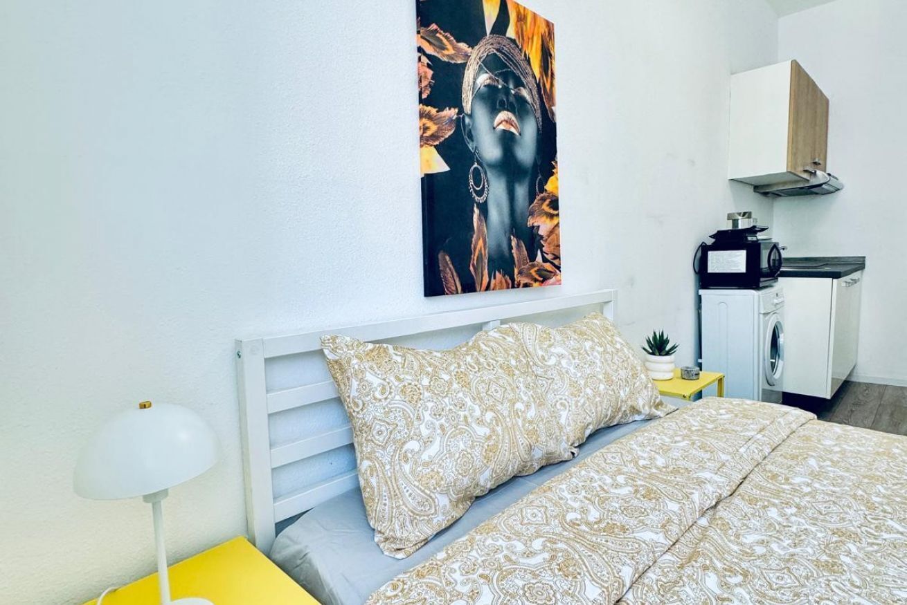 A bedroom with a bed, a nightstand, a lamp, a painting, and a kitchenette area.