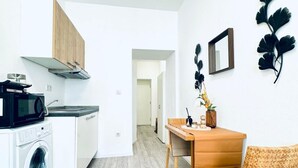 A compact kitchen with a microwave, washing machine, and wooden cabinets. A dining area with a table and chairs is visible, along with a mirror and wall decorations. - Comfortable and Affordable Palace Studio (Vienna)