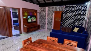 A modern living room with a wooden dining table, blue sofa, and a TV mounted on a wooden cabinet. - Encanto Farmstay Resort (Hunsur)