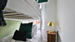 Una cama litera con ropa de cama blanca, un almohadón verde y una lámpara dorada sobre una mesita de madera.