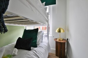 Un lit superposé avec une literie blanche, un oreiller vert et une lampe dorée posée sur une table de chevet en bois.