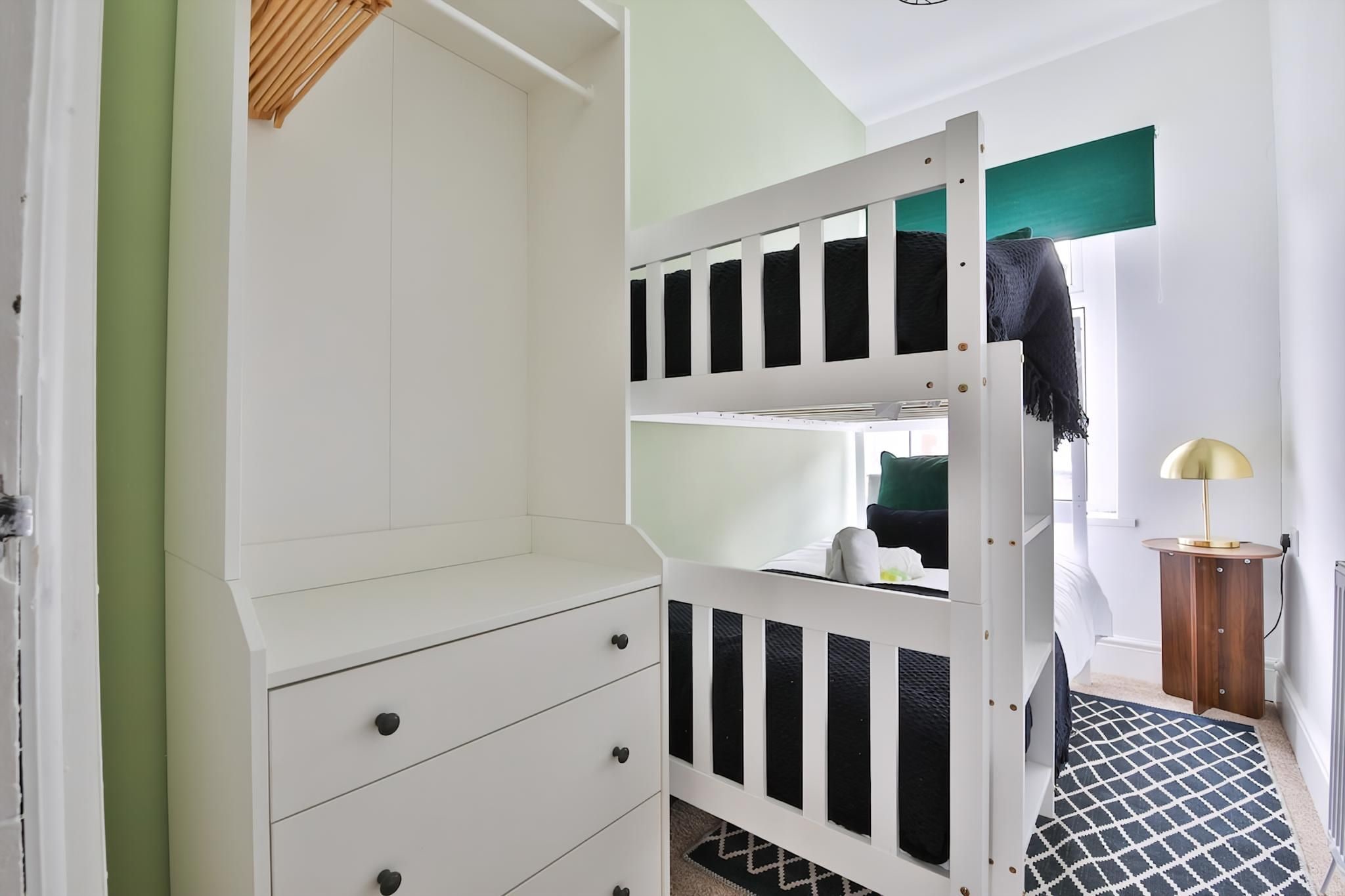 Uma cama beliche com estrutura branca, uma mesinha de madeira e um tapete estampado.