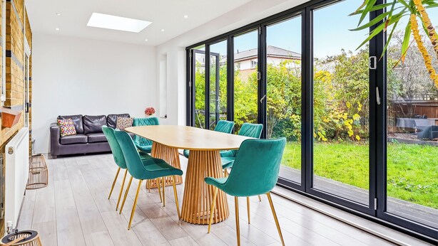 Un espace repas moderne avec une table en bois et des chaises de couleur turquoise, un canapé avec des coussins et de grandes fenêtres donnant sur un jardin et les maisons voisines.