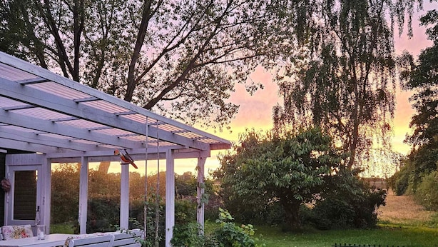 Veranda coperta con panchina e tavolo, circondata da vegetazione e un recinto.