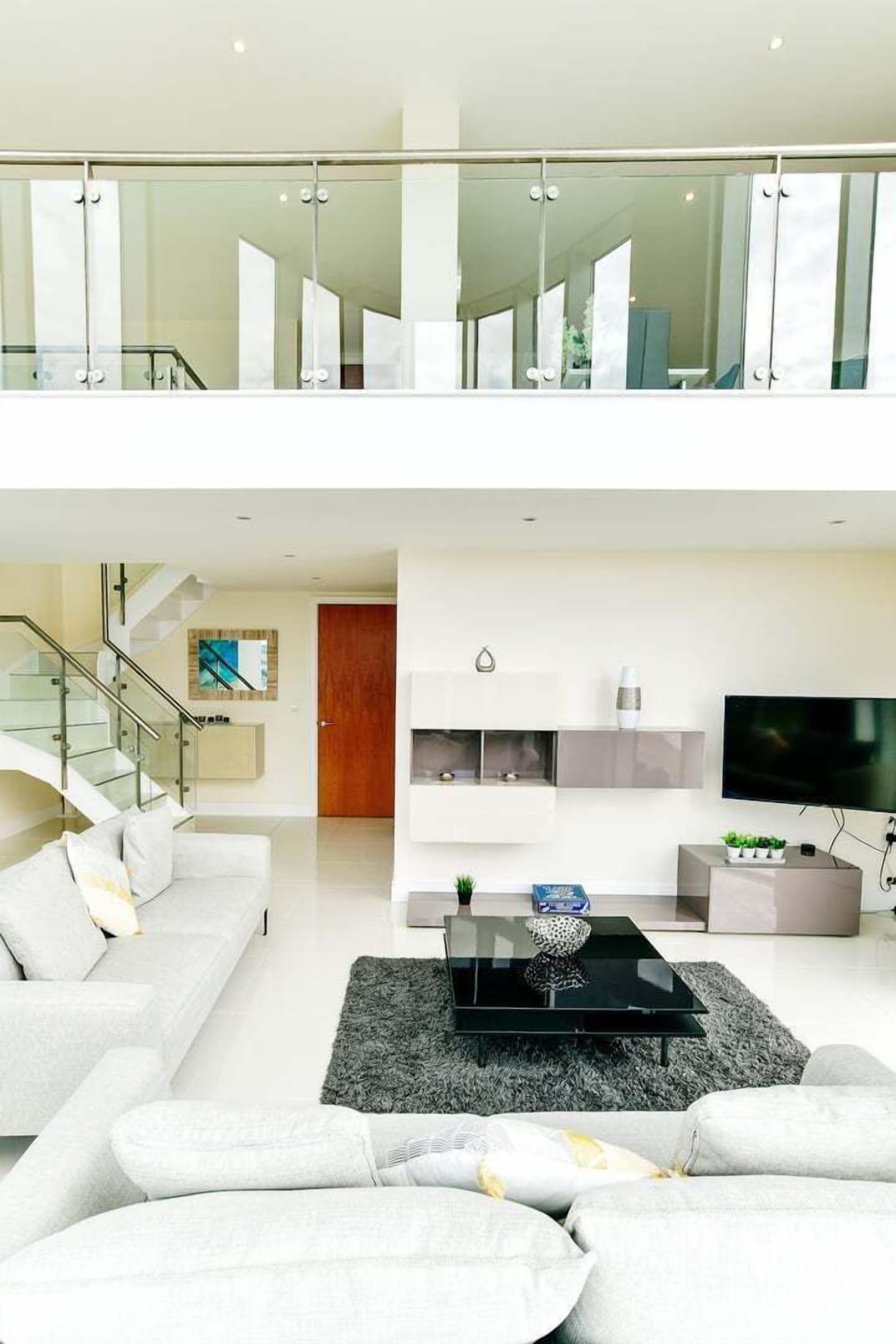 A modern living room with a sofa, a coffee table, and a TV. There is a staircase leading to an upper level with glass railings.