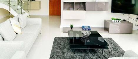 A modern living room with a sofa, a coffee table, and a TV. There is a staircase leading to an upper level with glass railings.