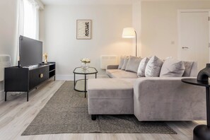 A modern living room with a grey sofa, a flat-screen TV, a small side table with a vase, and a lamp. - Toast Stays - The Elm at Parsons House (Washington)