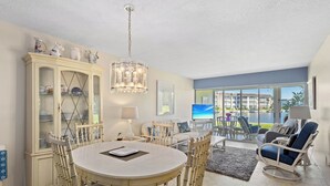 A dining area with a round table, chairs, a cabinet with glass doors, and a view of a pool and buildings through large windows.