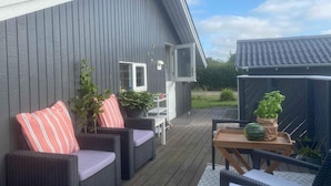 A covered outdoor patio with wicker furniture, a table with a watermelon, and a grey building with a window and a door. - 4 Star Holiday Home in Skaelskor (Skælskør)