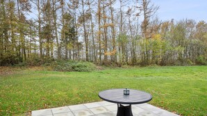 Ein runder Gartentisch mit Metallgestell auf einer Betonplatte, umgeben von einer Rasenfläche und einem bewaldeten Bereich.