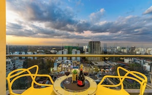 A balcony with yellow chairs, a table with two wine glasses and a bottle, and a city view under a cloudy sky.