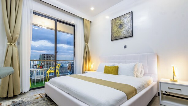 A modern hotel room with a large bed, a view of the city through floor-to-ceiling windows, and a balcony with outdoor seating. - Chic One Bedroom with Swimming Pool, Gym and magnificent City Views (Nairobi)