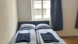 Un letto singolo con lenzuola a righe, una finestra con tende e vista sugli edifici.