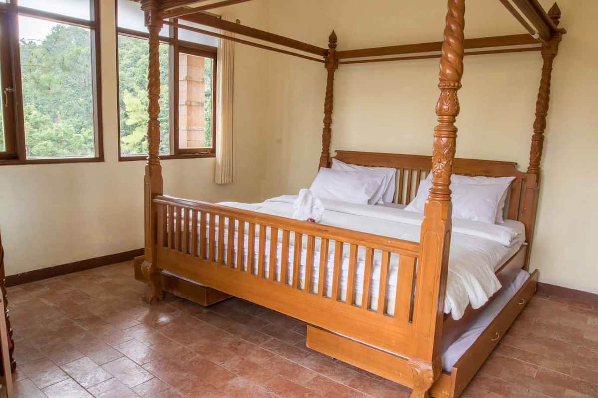 Uma cama de dossel com estrutura de madeira e roupa de cama branca em um quarto com piso de azulejos e uma janela com vista para vegetação.