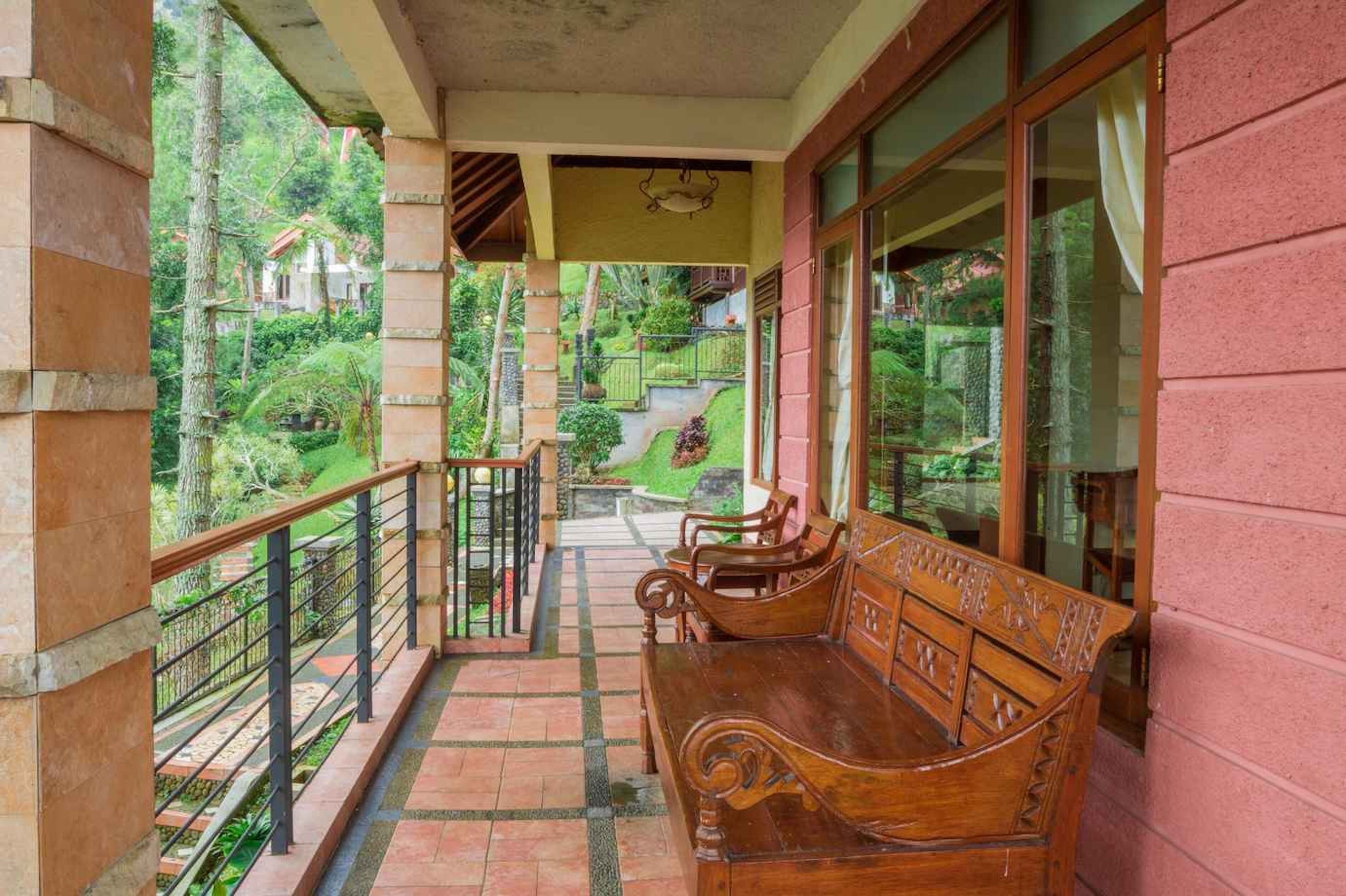 Un balcón techado con un banco de madera, barandilla y vistas a un jardín y edificios al fondo.