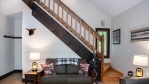 Una sala de estar con una escalera de madera, un sofá con cojines decorativos y dos lámparas sobre mesitas.