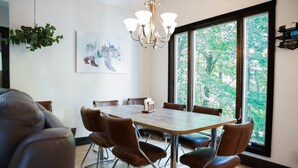 Comedor con una mesa grande, sillas de cuero marrón, una lámpara de araña y una ventana con vista a árboles.