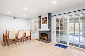 A modern living room with a fireplace, a dining area with wooden chairs, and a large sliding glass door leading to an outdoor area. - Near ONT Ontario Oasis Designer Home w Garden Park (Ontario)