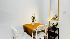 A small dining area with a table, two chairs, a potted plant, a desk with a lamp, and a mirror on the wall.