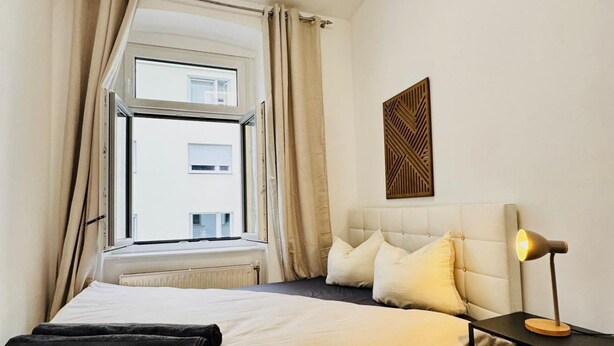 A hotel room with a bed, a nightstand with a lamp, a window with curtains, and a wall-mounted decor piece.