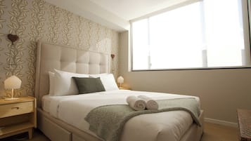 A neatly made bed with white linens and a green throw, a bedside lamp, and a nightstand with a lamp.